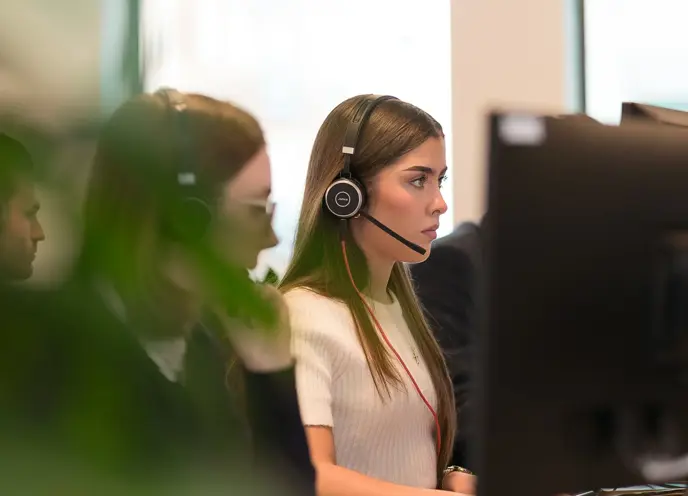 A woman wearing headset and sitting at a desk with other people in a modern office.
